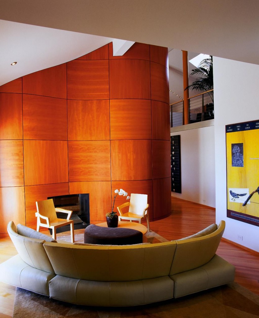 Modern semi-circular sectional couch wraps around yin-yang table in this living room flush with cherry wood, on flooring and large wave-shaped panel wall.