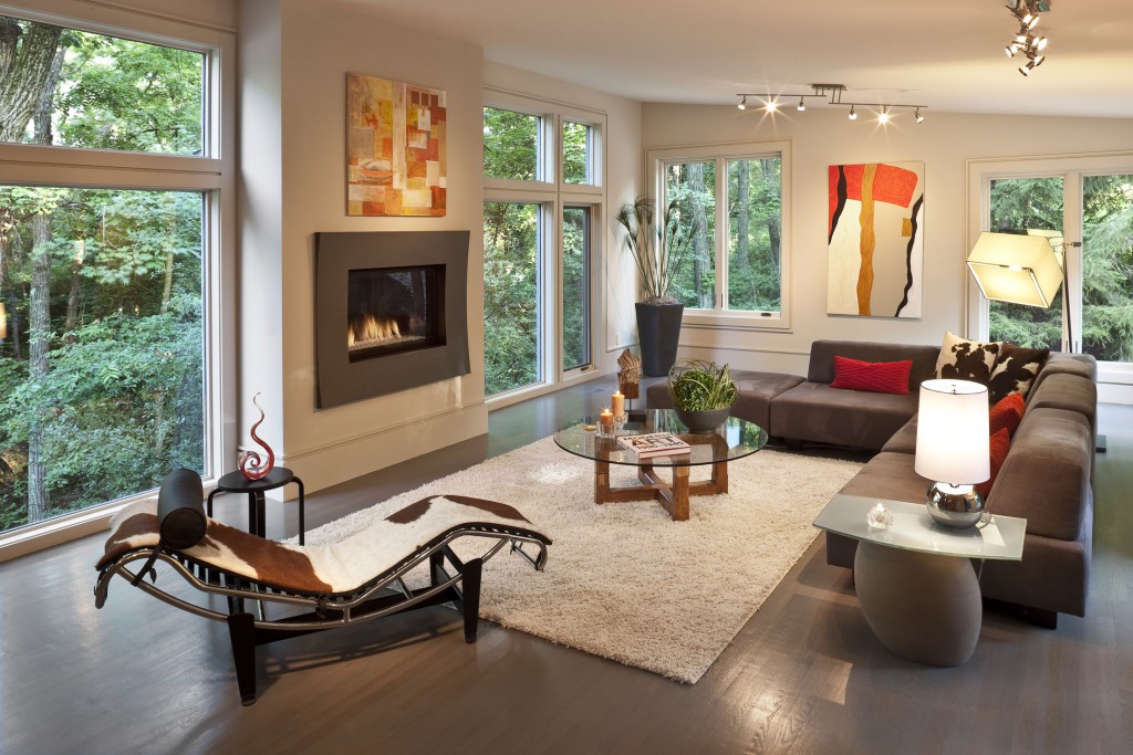 Sloped white ceiling over dark wood flooring in this living room holding modern armless brown sofa with red and cow-pattern pillows. White shag rug at center olds wood and glass coffee table, while cowhide chaise lounge sits on left.