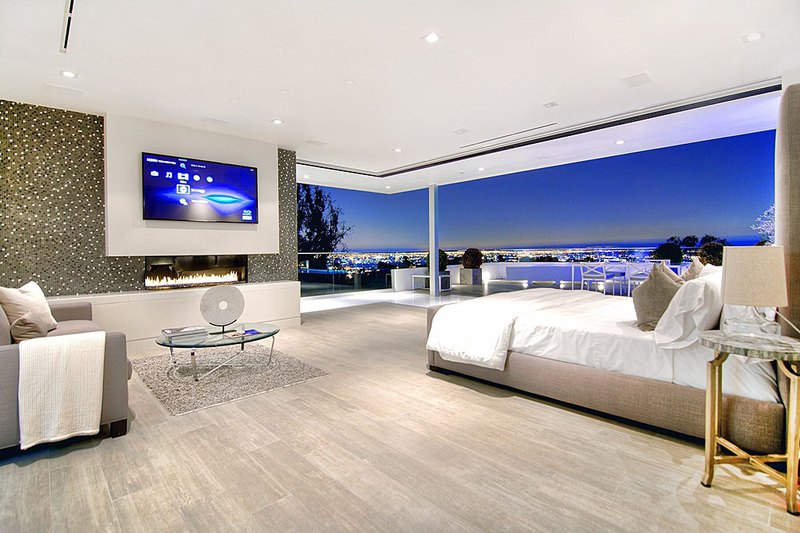 Modern spacious master bedroom design with extensive city views, floor-to-ceiling windows, small sitting area and large screen television mounted above a small gas fireplace