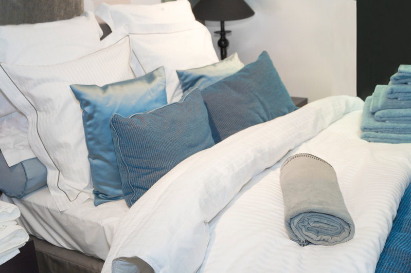 his pillow arrangement features beautiful blue pillows against delicately trimmed and subtly striped accent pillows matching the linens. A rolled accent pillow can be seen propping up the arrangement, while the sheen of peacock-blue catches the eye.