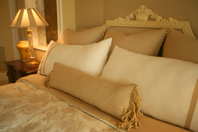 The gold and ivory design scheme of this room and the intricate attention to detail make this space exceptionally luxurious. An oblong gold throw pillow with a soft fringe sits against ivory and gold trimmed pillows and large square accent pillows in the rear. An opulent headboard acts as a frame, while the delicate gold and cream duvet cover ties everything together.
