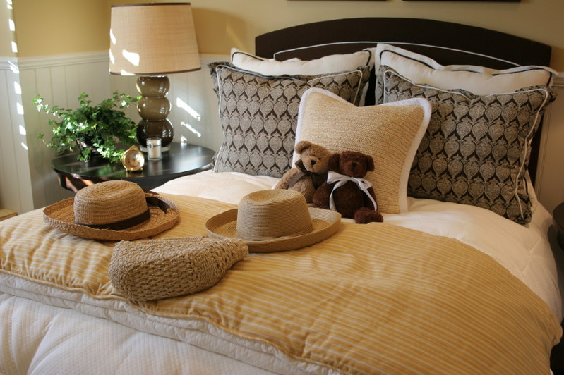 This charming bedroom showcases a variety of colors, patterns and textures. The natural fiber square accent pillow supports two adorable teddy bears, and goes perfectly with a variety of straw decorative accents. The darker rectangular throw pillows behind it offer a spectacular contrast against the lighter bed pillows that they rest against, while the dark headboard provides additional richness and beauty.