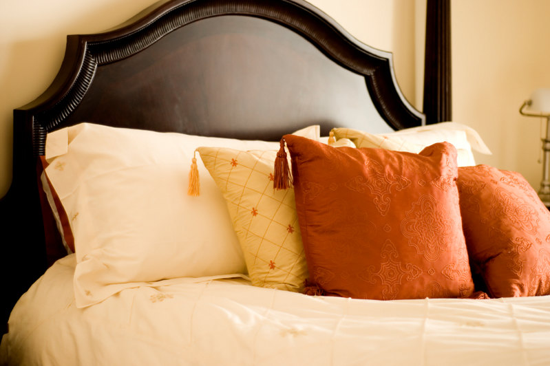 Rust orange and yellow pillows with corner tassels rest against the neutrality of a delicately embroidered cream bed pillow. A dark headboard offers optimal contrast for additional beauty and interest.