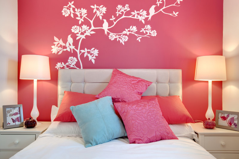 Stunning wall art steals the show in this bold, vibrant space. The art stands out in stark white contrast to the vibrant pink of the wall. A beige headboard complements the matching end tables and lamps, while additional pink, fuchsia, and blue pillows are tossed playfully upon the bed.