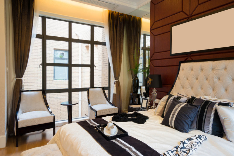 This outstanding bedroom features a luxurious ivory padded headboard against a chocolate backdrop. Black, white, and shimmering gray throw pillows mimic the colors seen in the stunning accent chairs, as well as the black trim of the windows overlooking an ivory brick wall outside.