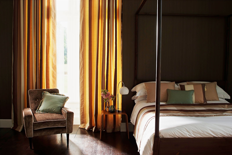 This bedroom features exquisite gold drapery that glows with the natural light filtering through the fabric. A soft armchair sits beside the four post bed, which is splendidly bedecked in complementary gold, green, ivory, and brown tones which tie the entire space together to stunning effect.