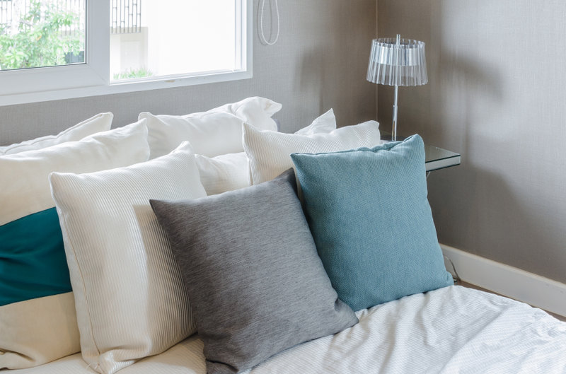 White accent pillows support a soft gray and blue pair of pillows upon soft bedding. Gray walls offer a fine canvas, and a delicate glass lamp brings additional interest to this quiet space.