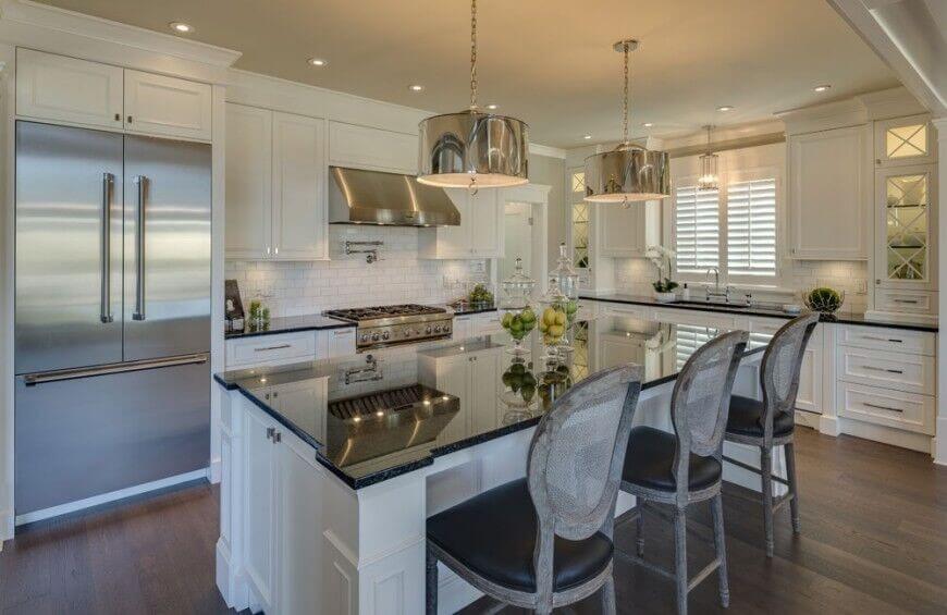 This beautiful kitchen makes great use of reflective surfaces to add interest and brighten the room. The dark counters and barstools bring weight to this broad room and complement the striking wood floor. The white cabinetry and tile backsplash keep the room feeling open and airy.